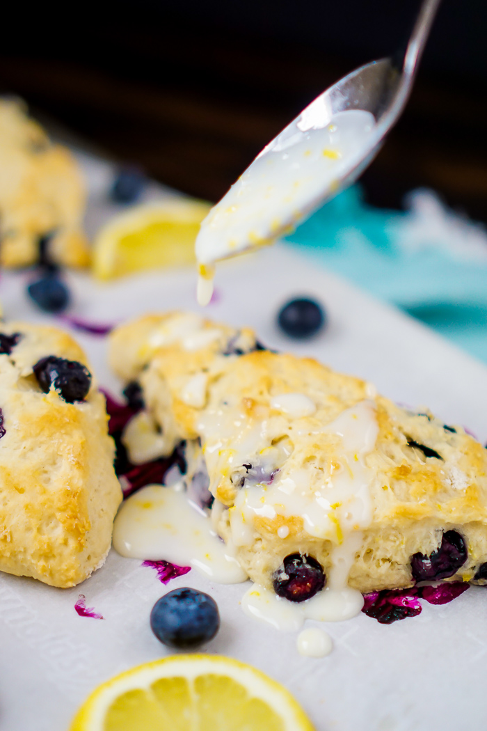 Three Blueberry and Lemon scones baked into triangle form are sitting on a piece of parchment paper with a spoon overhead drizzling on a citrus lemon glaze with lemon zest in it. 