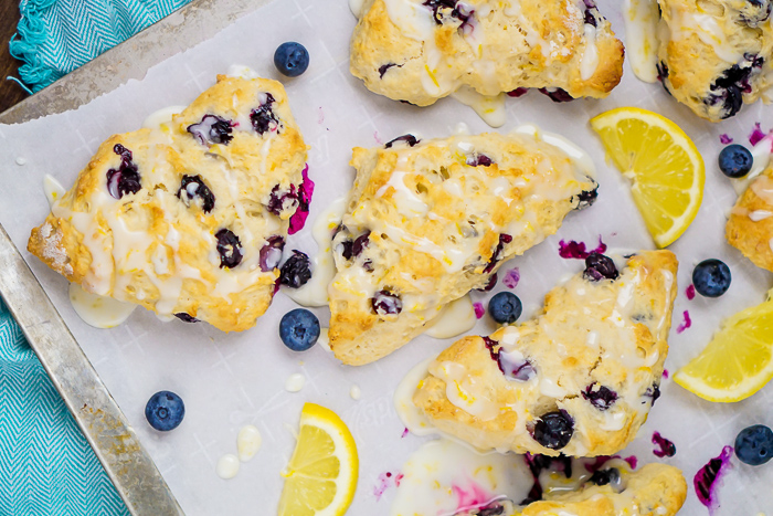 Lemon and Blueberry Scones in triangle shape sitting on a piece of parchment paper on a baking sheet with lemons and blueberries sitting around the scones loose on the pan