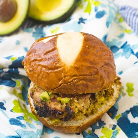 Turkey burger with chopped avocado inside sits on a pretzel bun on top of a blue and green floral napkin with extra avocado sits in the back left corner