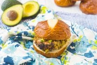 Turkey burger with chopped avocado inside sits on a pretzel bun on top of a blue and green floral napkin with extra avocado sits in the back left corner