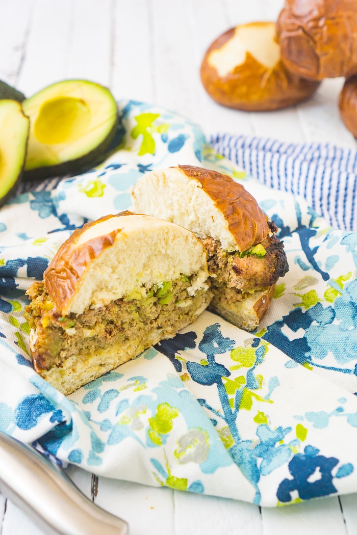 Ground turkey and avocado burger is cut open to show chunks of avocado inside the burger while it sits on a pretzel bun on top of a blue and green floral napkin with extra avocado sits in the back left corner