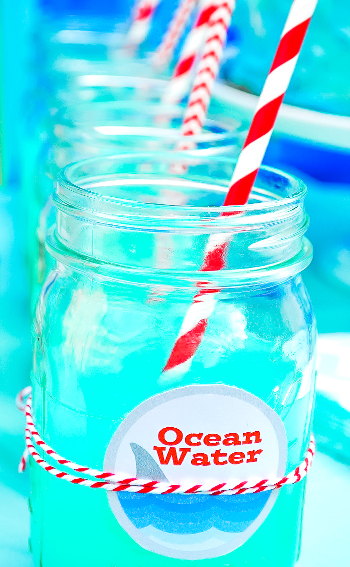 A row of mason jars filled with a blue punch and topped off with a logo on the front that says Ocean Water and a red and white striped straw resting in the jar