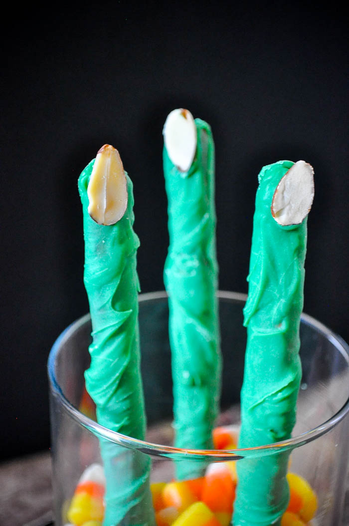 3 pretzel rods decorated as Frankenstein fingers with green candy melt and almond slice fingers nails stand up in a glass jar with candy corn at the bottom. 