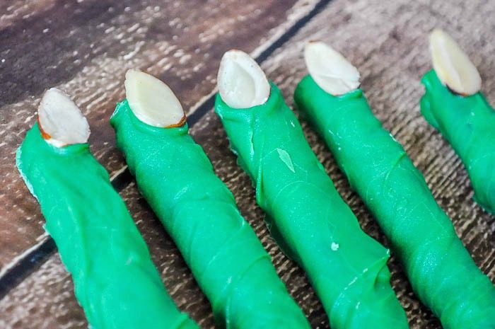 5 Pretzel rods sit in a row on a dark wood table and are dipped in green candy coating with an almond slice as the finger nail so it looks like Frankenstein Monster's Fingers