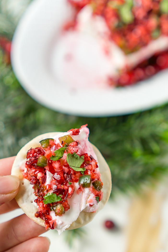 cranberry and jalapeno dip with cream cheese spread is on top of a round cracker with a plate of the dip in the background out of focus