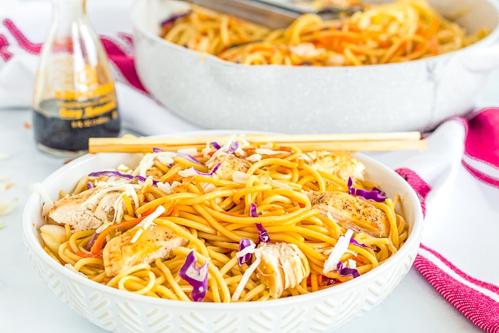 A big white pasta bowl is filled with chicken lo mein with cabbage, carrots and onions with a set of chopsticks rest on the side of the bowl with a skillet full of lo mein sitting in the background out of focus