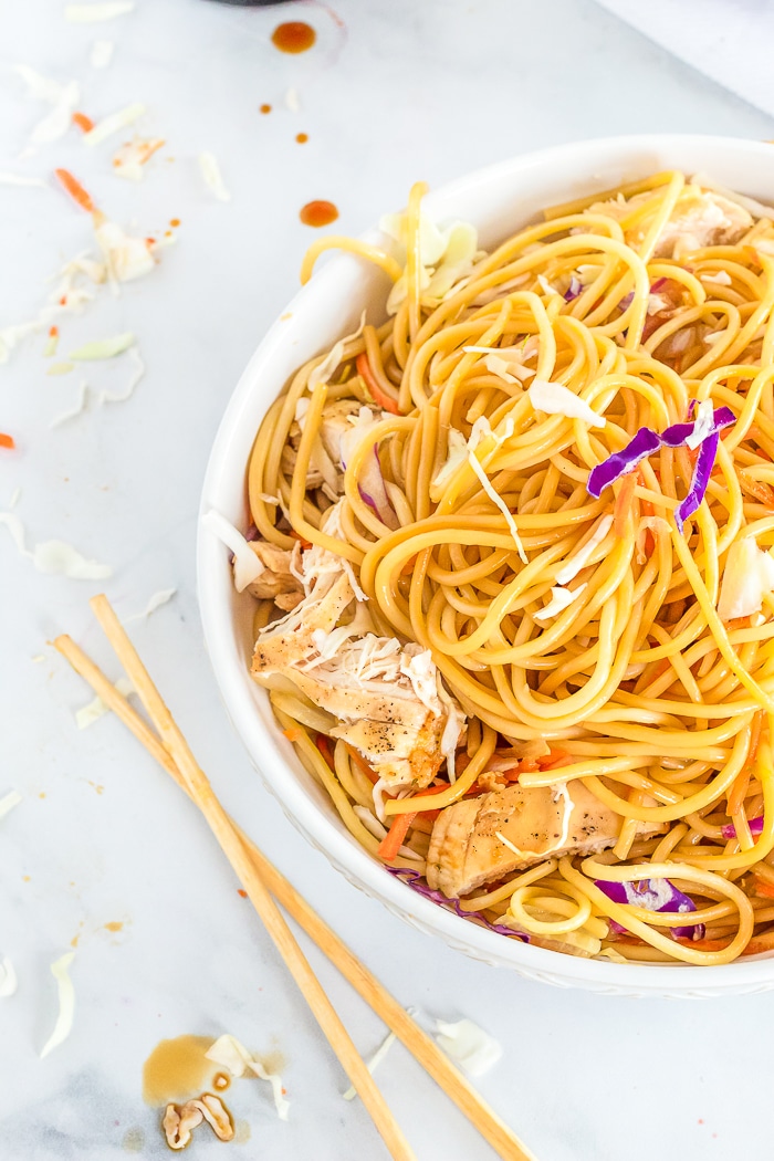 Straight down shot onto a round white bowl of chicken lo mein with purple cabbage, carrots and onions. A set of chopsticks sits next to the bowl on the white marble countertop with a little cooking mess surrounding them.