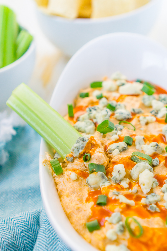 An oval white baking dish is filled with orange buffalo chicken dip, blue cheese crumbles and chopped green onions with a celery stick sticking out of the dish on the left hand side. A white bowl of celery sticks and a white bowl of tortilla chips are slightly out of frame in the top left corner.
