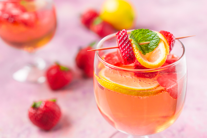 Strawberry Rosé Spritzer in a low stemmed wine glass garnished with fresh strawberries, a lemon slice, and a mint leaf. Fresh strawberries and lemons sit on the pink countertop around the glass. 