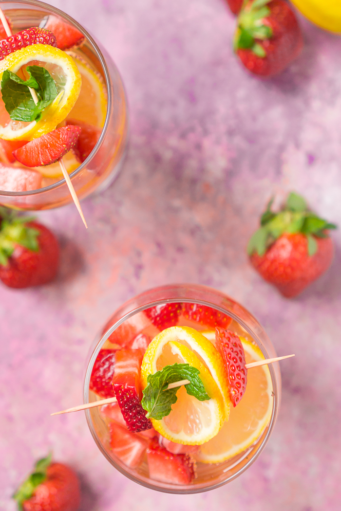 Straight down shot of two wine glasses on a pink tabletop filled with wine spritzer recipe using rosé wine, fresh strawberries, lemons, and a mint garnish. 