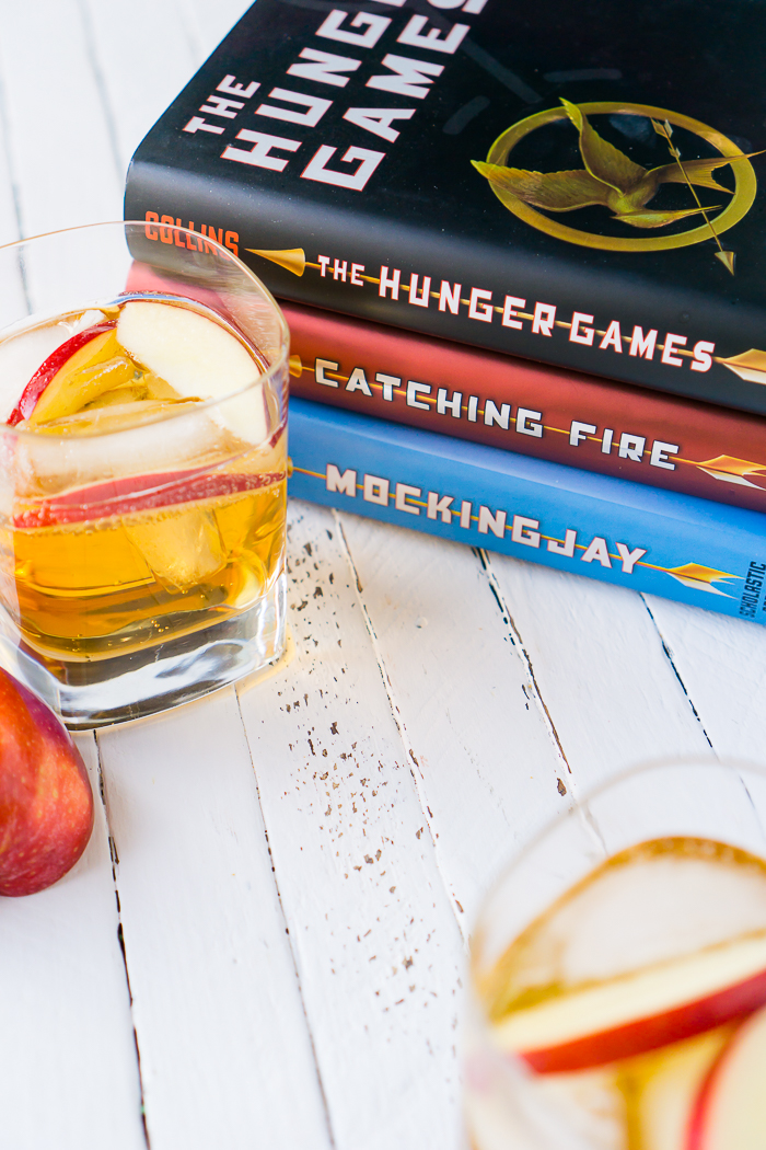 All three hunger games books sits in a stack on a white wood tabletop next to a hunger games inspired fireball cocktail with fireball whisky and hard apple cider named Girl on Fireball that is garnished with red apple slices