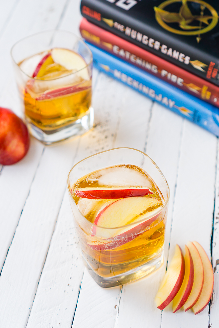 Fireball Whisky mixed with angry orchard hard cider sits in a square old fashioned glass garnished with apple slices and sitting in front of the Hunger Game Books