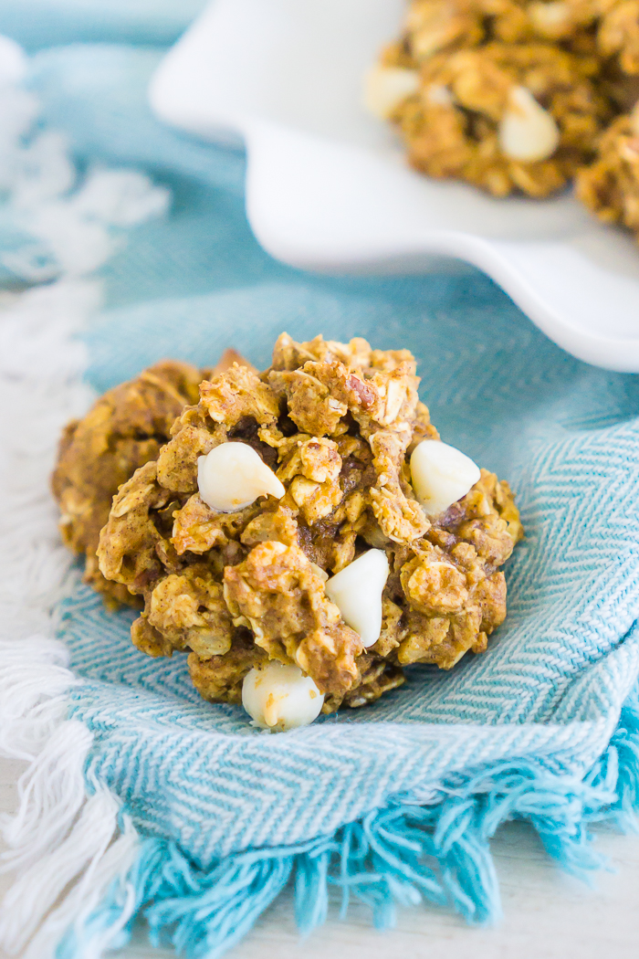 A baked pumpkin oatmeal cookies with white chocolate chips and chopped pecans sits on an aqua blue napkin with white fringe with a white plate with more cookies sits slightly out of focus on the top right corner. 