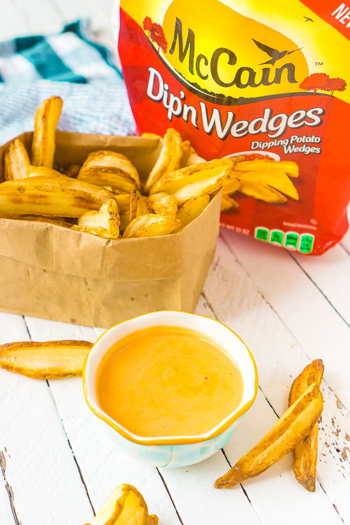 White and aqua bowl with an orange dipping sauce inside sitting in front of a paper bag folded down with fries inside and a red and yellow McCain Dip'n Wedges bag 
