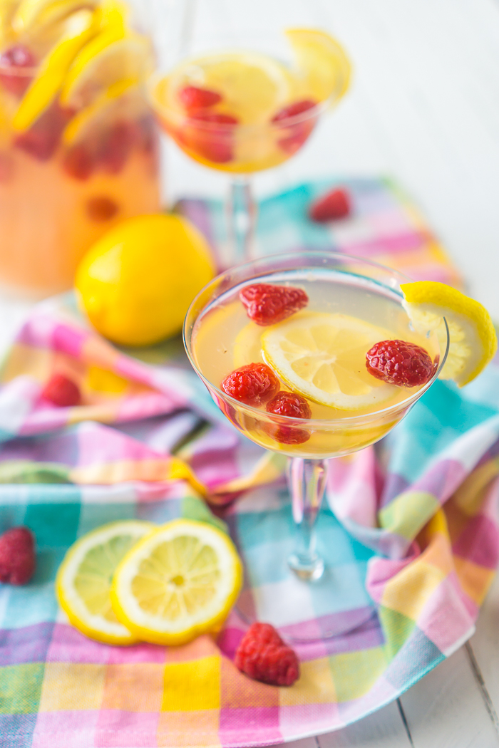 Two champagne coup glasses filled with raspberry lemonade sangria, slices of lemons, and raspberries. Pitcher of sangria is tucked back in the top left corner and a bright blue, pink and purple plaid napkin sits under the glasses on a white table. 
