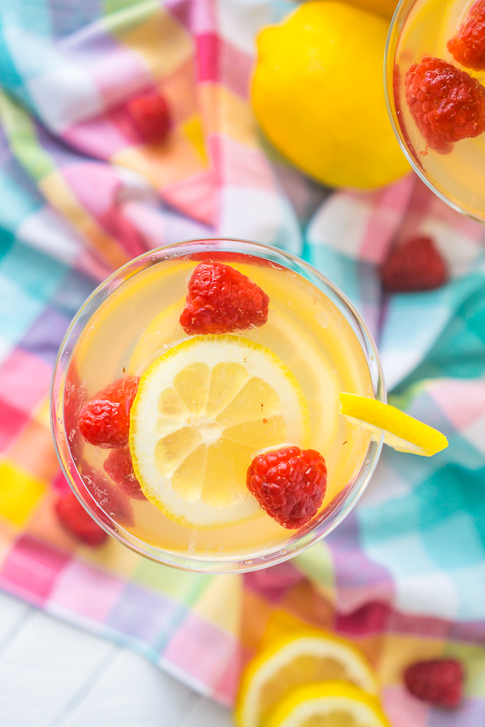 Straight down shot onto a coupe glass with lemon slices, fresh raspberries and white wine lemonade sangria sitting on a bright blue, purple, pink and yellow plaid napkin. 