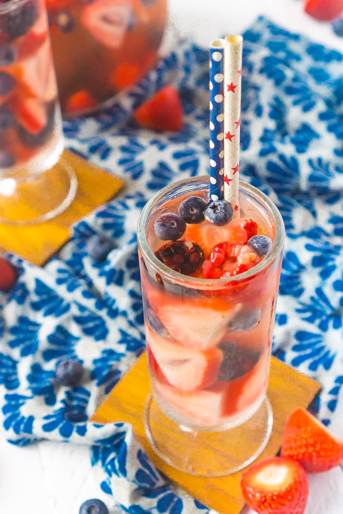 A red white and blue sangria sits in a tall, round glass with white wine and red and blue summer berries. A blue polka dot straw and white straw with red stars sits inside the cocktail with a second glass and the sangria pitcher sitting off to the side. 