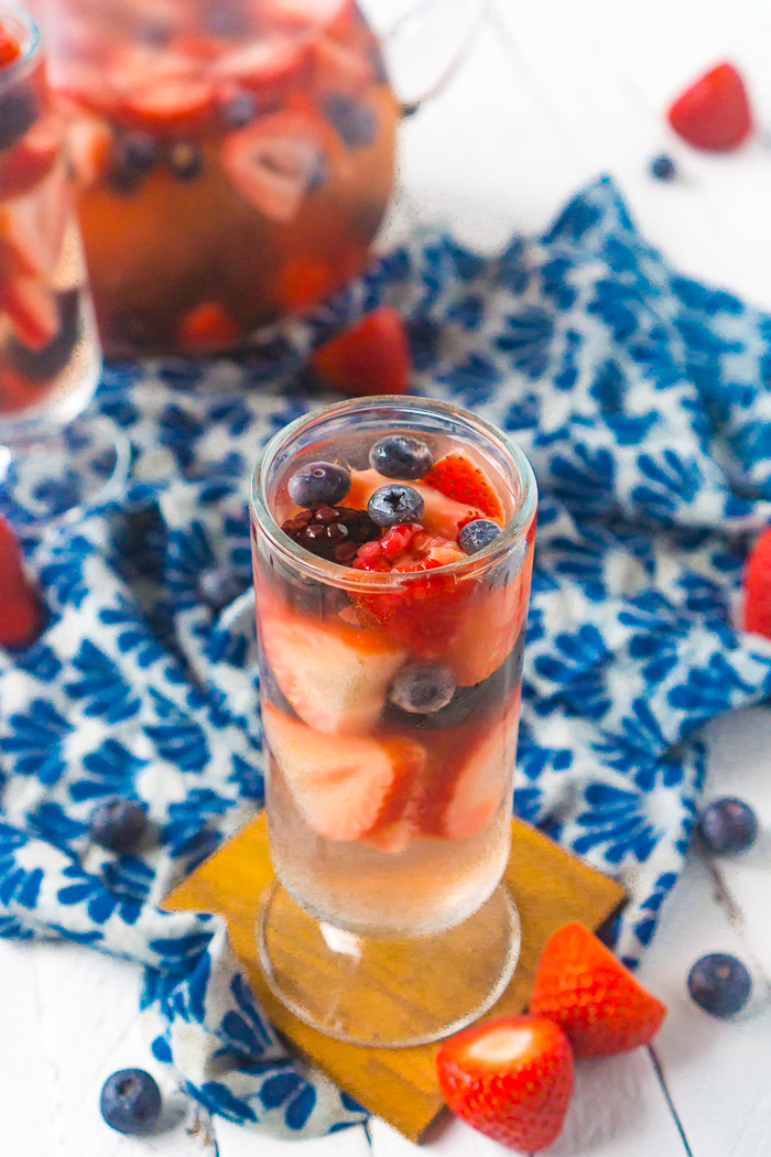 Tall, round cocktail glass filled with white wine sangria and strawberries, blueberries, blackberries and raspberries sits on top of a square wood coaster. The sangria pitcher sits in the back left corner partially out of sight, and extra berries are sitting around. 