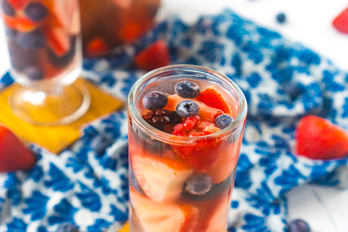Tall, round cocktail glass filled with white wine sangria and strawberries, blueberries, blackberries and raspberries. Another glass and the pitcher sits in the back left corner partially out of sight. 