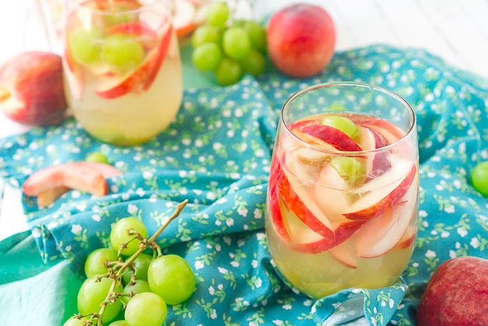 Two stemless wine glasses filled with white wine sangria with slices of white peaches and green grapes sitting on a blue napkin with white flowers. Peaches and stemmed grape bundles sit scattered around the glasses. 