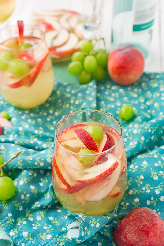 A stemless wine glass with white peach sangria, white peach slices and halved green grapes sit on a flowered blue napkin that is sitting on front of a second glass, the sangria pitcher, an empty wine bottle as well as a whole peach and stemmed grapes out of focus. 