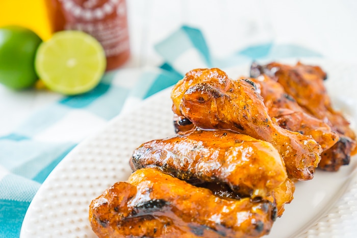 Plate of chicken wings tossed in a honey sriracha sauce placed on a blue plaid napkin with a jar of honey and a bottle of sriracha in the back left over. 