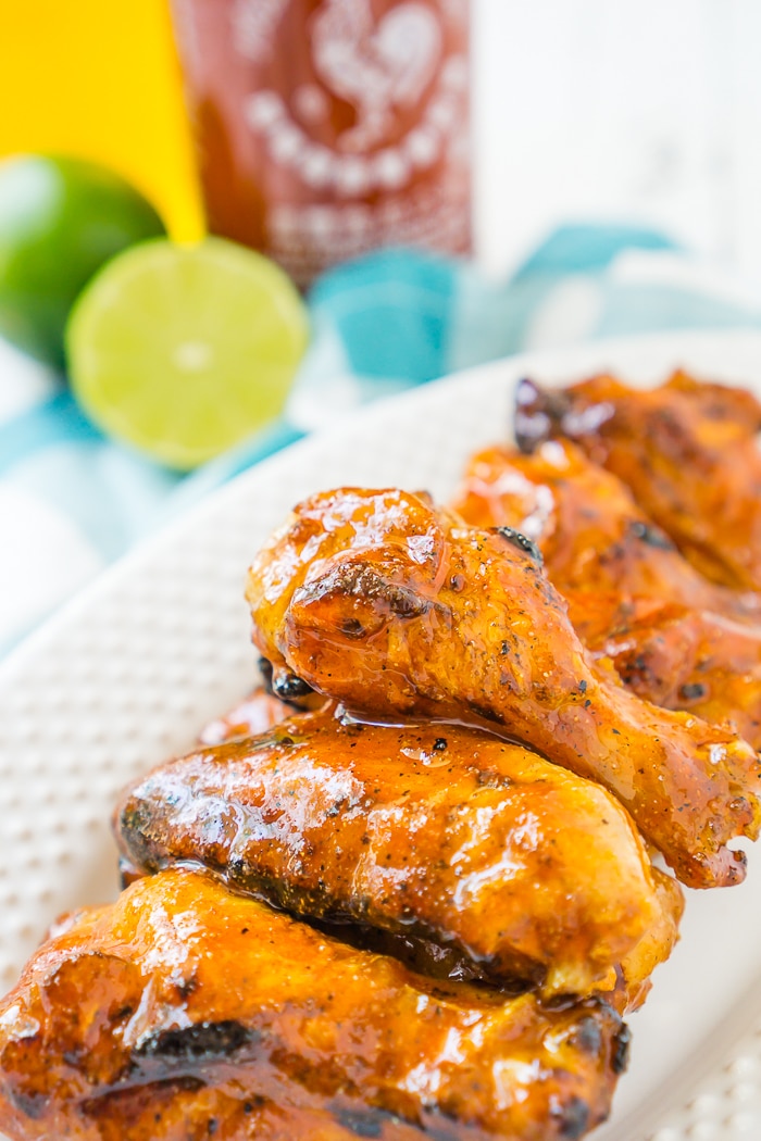 A white oval plate of 5 chicken wings tossed in a honey sriracha chicken wing sauce. Bottle of Sriracha sauce, a jar of honey and an open lime is out of focus in the top left corner. 
