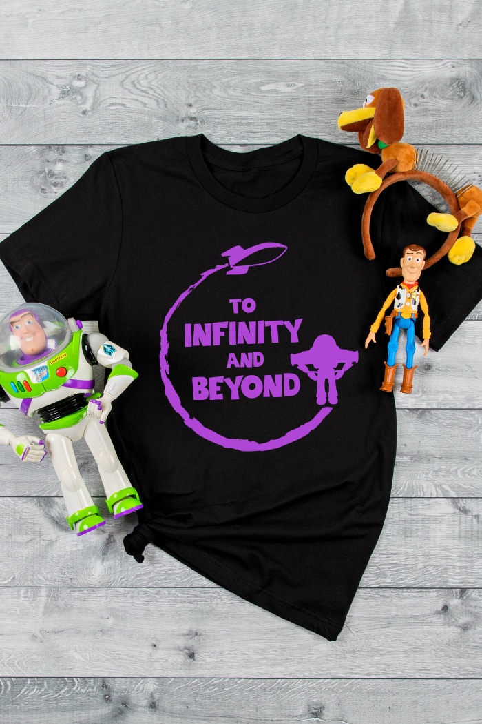 To Infinity and Beyond SVG File with Buzz standing wings out and a rocket ship blasting off is ironed onto black tshirt with purple heat transfer vinyl as a Disney World Shirt with Slinky Mickey Ears and Woody and Buzz action figures sitting by the shirt. 