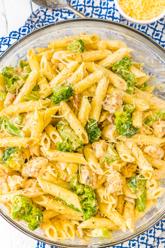 A straight down photo of a clear glass pyrex bowl sitting on a blue and white napkin that is filled with penne pasta, chicken, bacon, and broccoli florets coated with a lighter Alfredo sauce made with cauliflower. A bowl of shredded parmesan cheese sits in the top right corner of the photo. 
