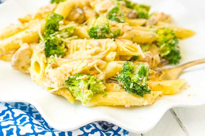 Up close photo of a wavy white plate sitting on a blue and white napkin topped with a serving of penne pasta with chicken, bacon, and broccoli that is coated with a lighter Alfredo sauce made with cauliflower. 