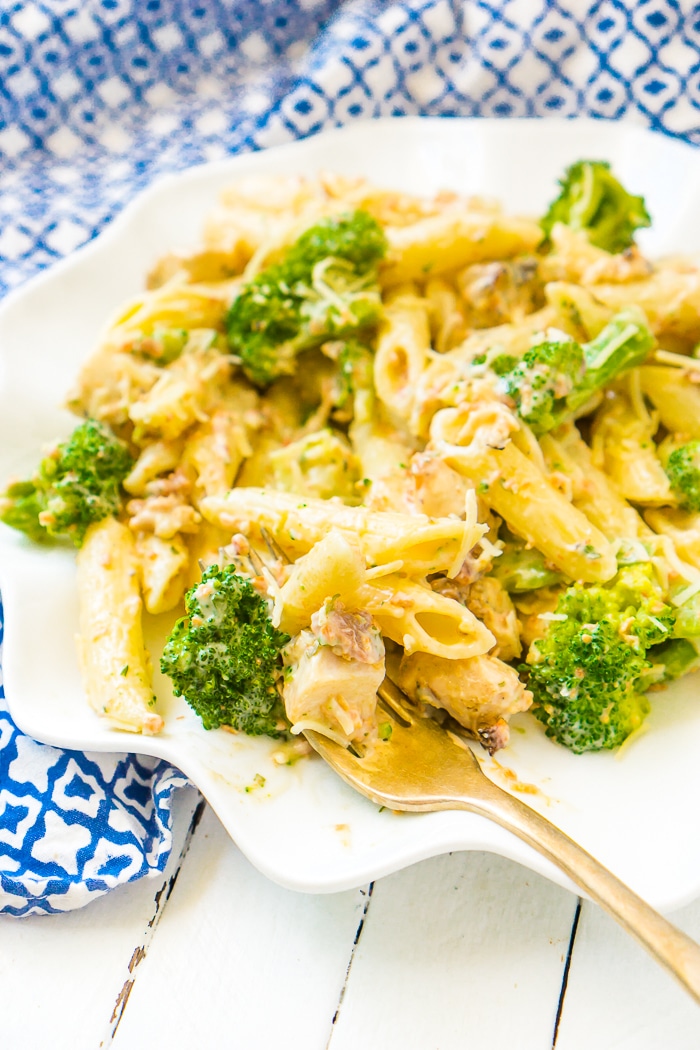 Up close photo of a wavy white plate sitting on a blue and white napkin topped with a serving of penne pasta with chicken, bacon, and broccoli that is coated with a lighter Alfredo sauce made with cauliflower. A fork rests on the bottom right of the plate with pasta and chicken resting on it.
