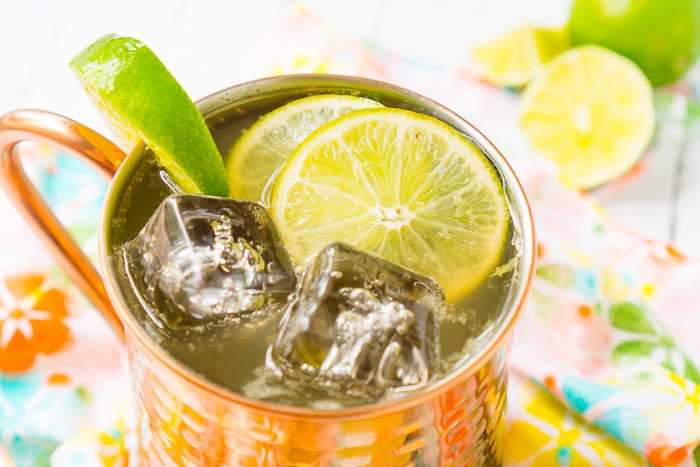 Up close photograph of a copper mug filled with a moscow mule with square ice cubes and lime slices at the top of the drink and a lime wedge on the edge of the mug. The mug sits on a coral floral napkin on a white table with lime wedges in the top right corner. 