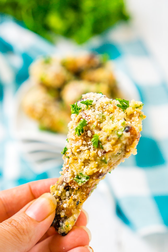 Up close photo of a chicken wing coated in garlic, parmesan and chopped parsley being held up with a blue plaid napkin in the background that has a plate of chicken wings and a bunch of fresh parlsey in the background out of focus