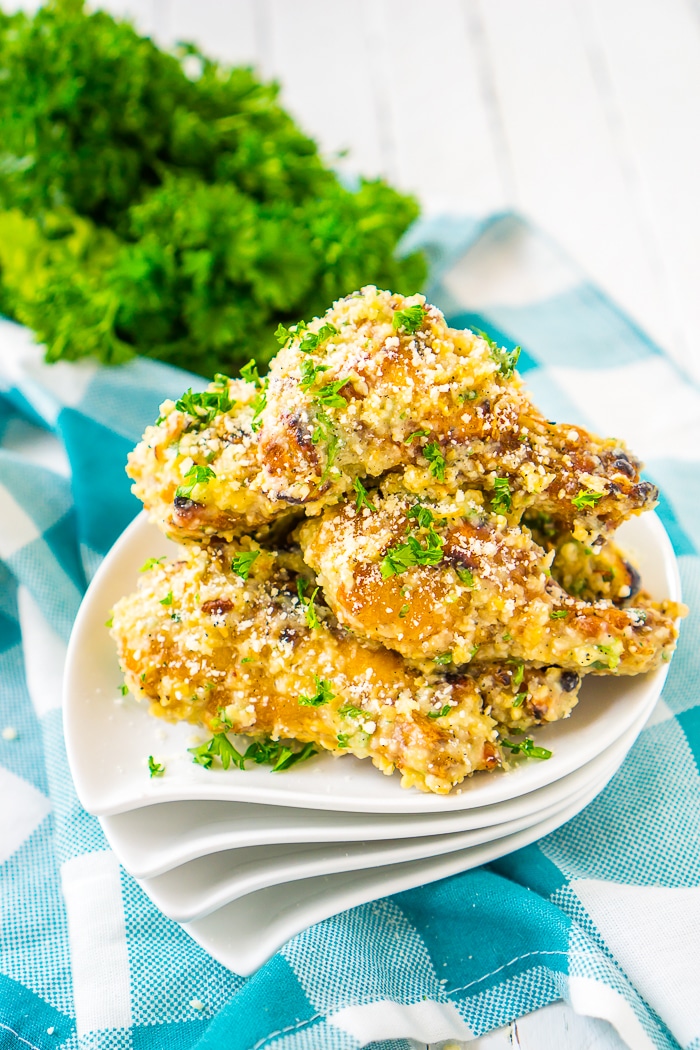 A stack of 4 round white appetizer plates sits on a blue plaid napkin and are topped with chicken wings tossed in a garlic parmesan chicken wing sauce. A bunch of fresh parsley sits in the top left corner. 