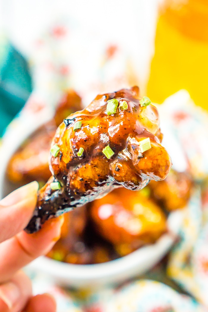 Up close photo of fingers holding a chicken wing smothered in a honey bbq sauce and garnished with little green onions. In the background is a white bowl filled with more chicken wings and a jar of yellow honey, all out of focus. 