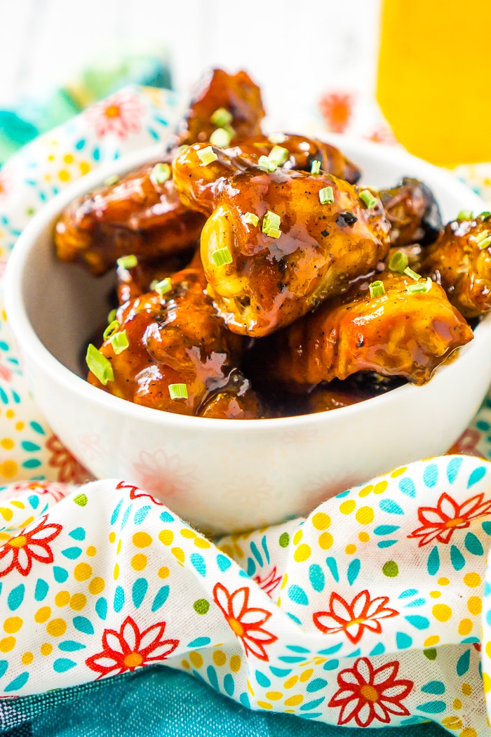 A small white bowl is sitting on an aqua, yellow and coral floral napkin filled with chicken wings smothered in a honey bbq chicken wing sauce and garnished with green onions. A jar of yellow honey sits to the top right out of focus. 