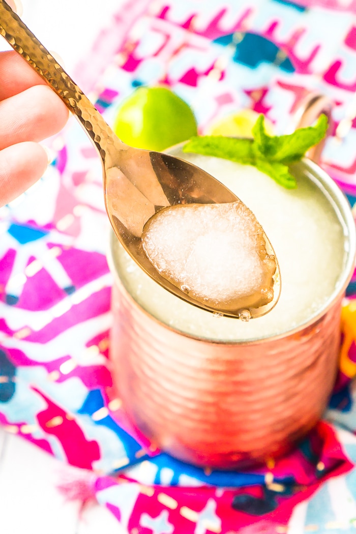 A copper spoon has frozen moscow mule slushie on it and is hovering over a copper mug filled with the slushie behind it on a dark pink napkin. 