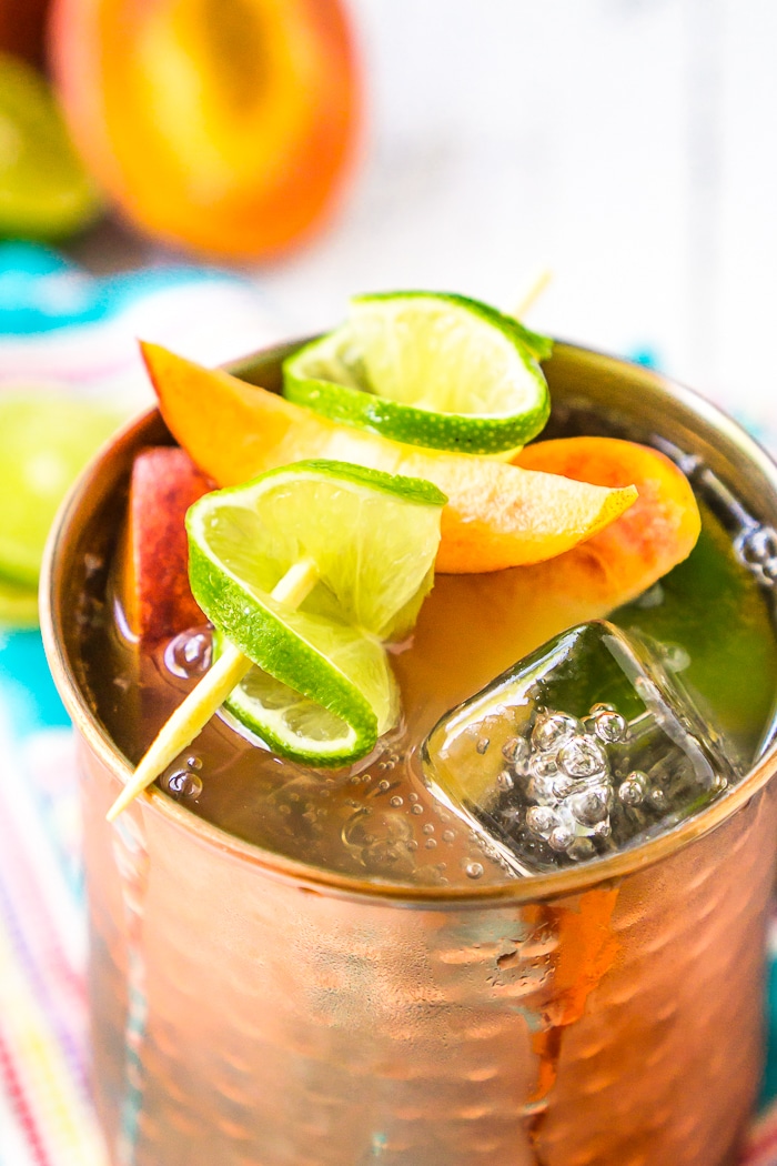 Copper mug is resting on top of a blue napkin and is filled with fresh peaches and lime slices on top of a peach Moscow mule. 