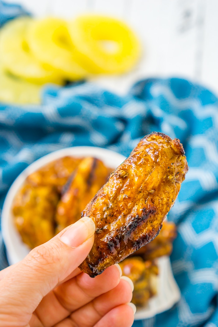Hand is holding up a grilled chicken wing tossed in a teriyaki and pineapple sauce with a plate of wings on the white wood table in the background as well as slices of pineapples. 