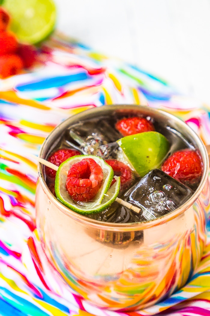 A copper mug sits on a bright colorful napkin and is filled with square ice cubes and raspberry Moscow mules with a raspberry and lime slice garnish resting on top. 