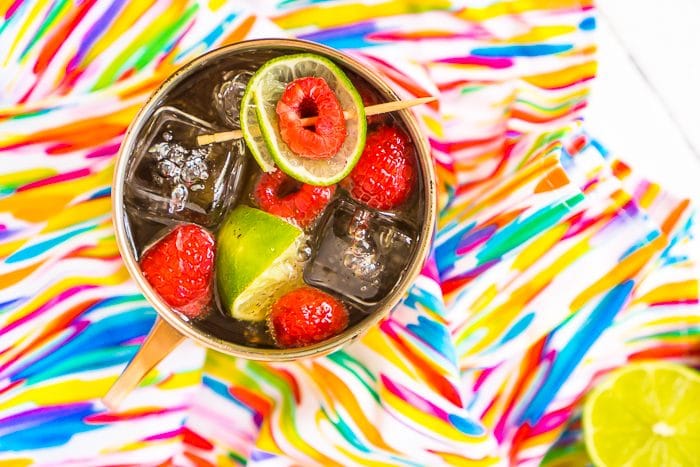 Straight down shot onto a copper mug filled with square ice cubes, raspberries, lime wedge and raspberry Moscow Mule resting on a bright and colorful paint splatter napkin. 