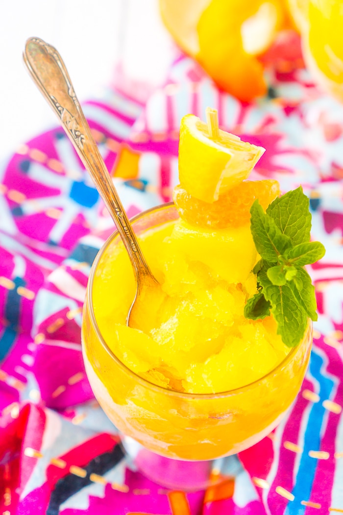 A close up photo of a round wine glass with yellow slushie and a garnishment of mint leaves, lemon wedge, orange slice and pineapple chunk with a spoon standing up inside the glass. 