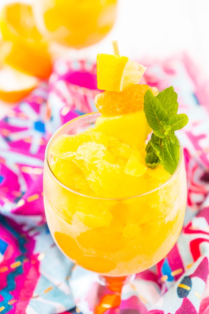 Short stemmed goblet is filled with a yellow tropical rum punch slushie and garnished with fresh mint and a stick with a lemon wedge, an orange slice, and a pineapple wedge. In the background is a second glass of alcoholic slushie next to fresh orange peels.  