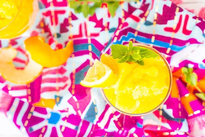 A straight down shot of a round wine glass filled with a yellow tropical rum punch alcoholic slushie topped with a garnish stick of lemon wedge, orange slice, and pineapple wedge plus a sprig of mint. In the background is a bright pink napkin and another glass is in the back left corner with an orange peel next to it. 