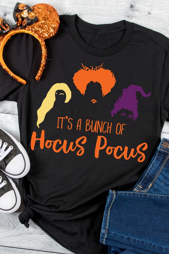 A black tshirt has a Halloween Hocus Pocus design on it from Heat transfer vinyl that shows yellow blonde hair, orange hair and purple hair to represent the Sanderson sisters from Hocus Pocus with orange words underneath that says It's a bunch of hocus pocus! also with the shirt is a pair of orange sequined mickey ears for halloween, black sneakers and jeans. 