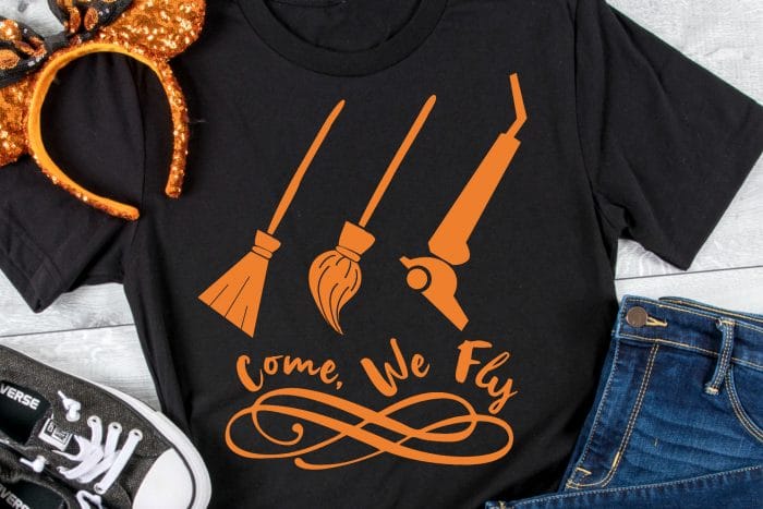 A black tshirt has an orange HTV design on it that says come we fly with a room, mop, and vacuum and it sits with orange sequined mickey ears for halloween, black sneakers and jeans. 