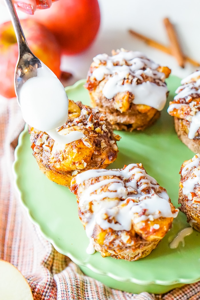 A spoon is drizzling white icing onto cinnamon roll muffins with chopped apples and pecans inside. 
