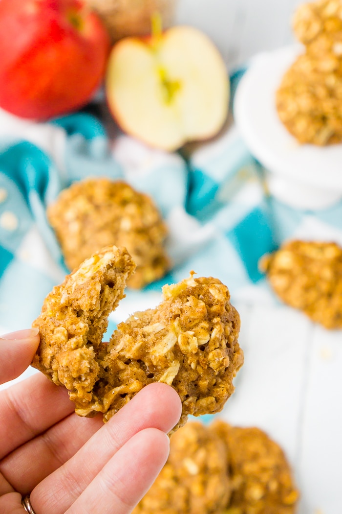 A HOMEMADE OATMEAL COOKIE WITH CHOPPED APPLES IN IT IS RIPPED IN TWO AND BEING HELD UP BY A HAND WHILE A BLUE PLAID NAPKIN, A CUT OPEN RED APPLE, AND OTHER COOKIES ARE BLURRED IN THE BACKGROUND ON A WHITE COUNTER TOP