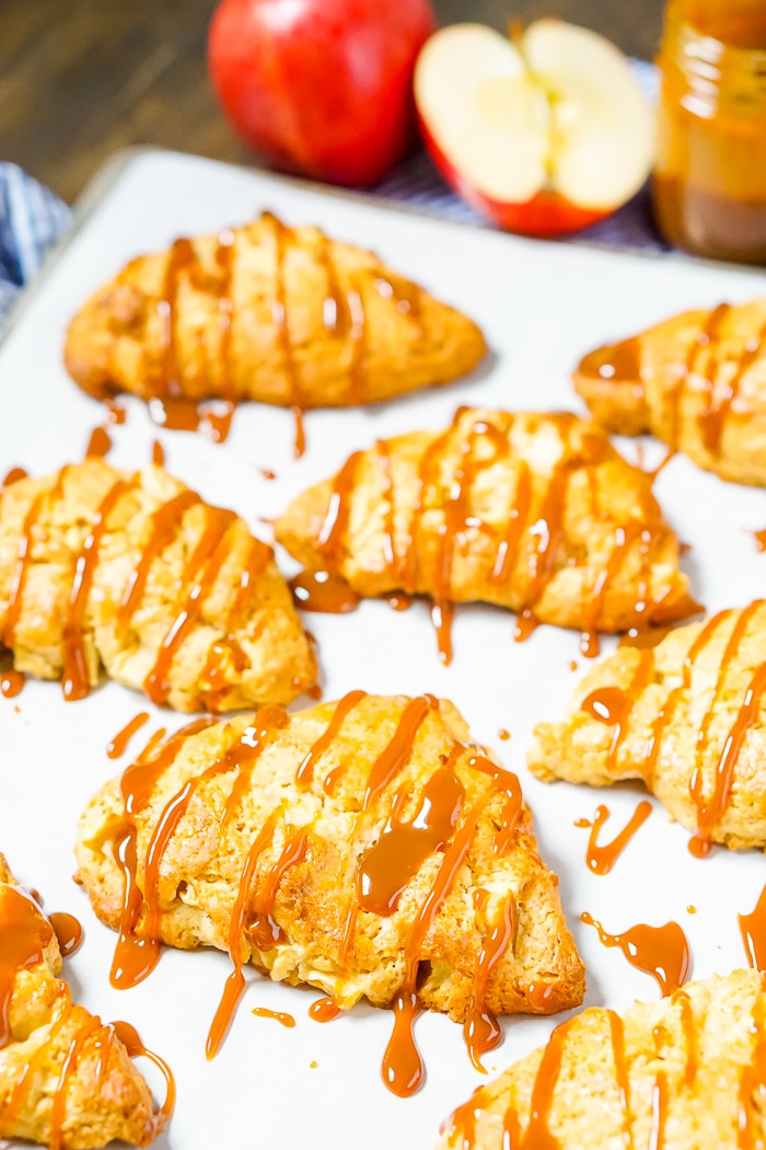 A baking sheet lined with parchment paper has 6 caramel apple scones on them with caramel drizzle on top and on the parchment pan. A bottle of caramel and a couple apples sit in the background. 