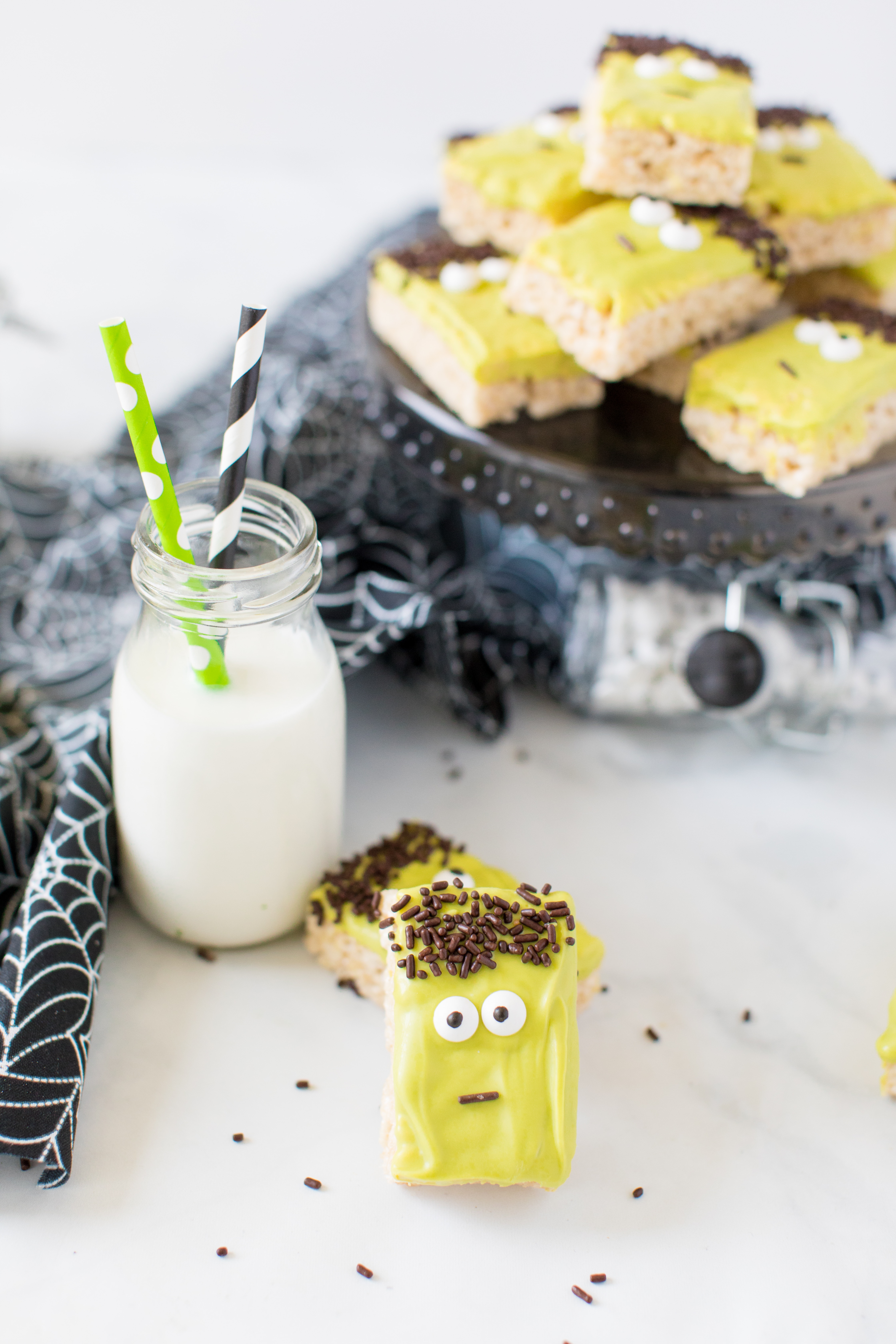two frankenstein rice krispie treats rest in front of a small milk jug with green and black paper straws in front of a black cake plate with more of the halloween treat